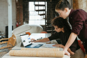 Man and woman at a desk looking at a Microsoft Surface. The woman is pointing at something on the Surface. Materials for refurbishing furniture can be seen in the foreground.