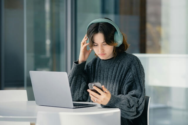 Woman sitting in front of a laptop, wearing headphones and looking at a smartphone