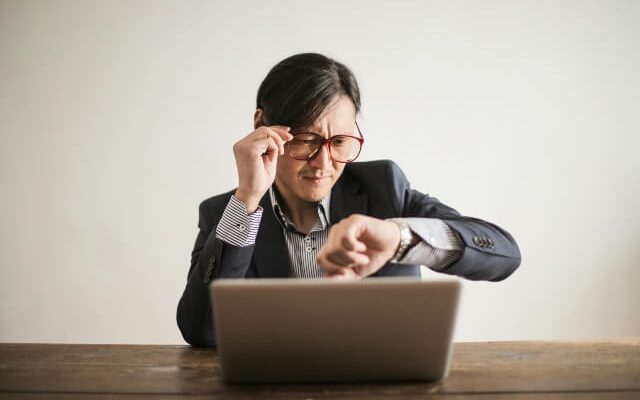 Man in business attire sitting in front of a laptop looking at his watch