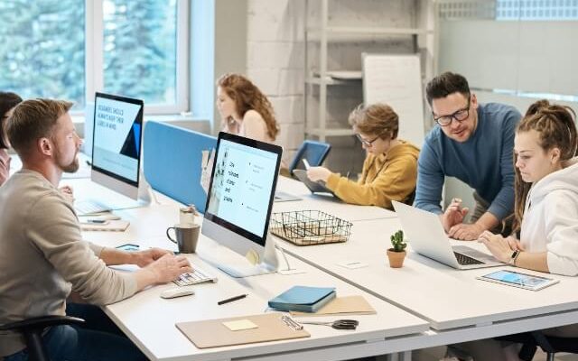 A group of employees working on desktop, laptop and tablet devices. A manager is looking at a laptop with one employee.