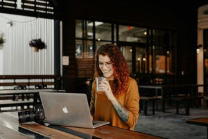 Woman sitting at a table with a laptop computer and digital camera. She is smiling and drinking iced coffee.