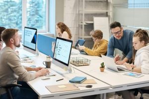 A group of employees working on desktop, laptop and tablet devices. A manager is looking at a laptop with one employee.